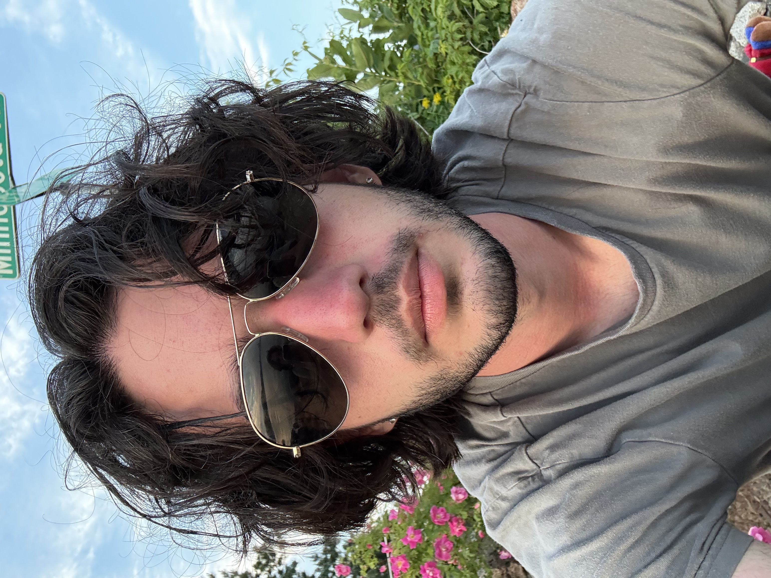 Maikol outdoors wearing sunglasses, with flowers and blue sky behind him.